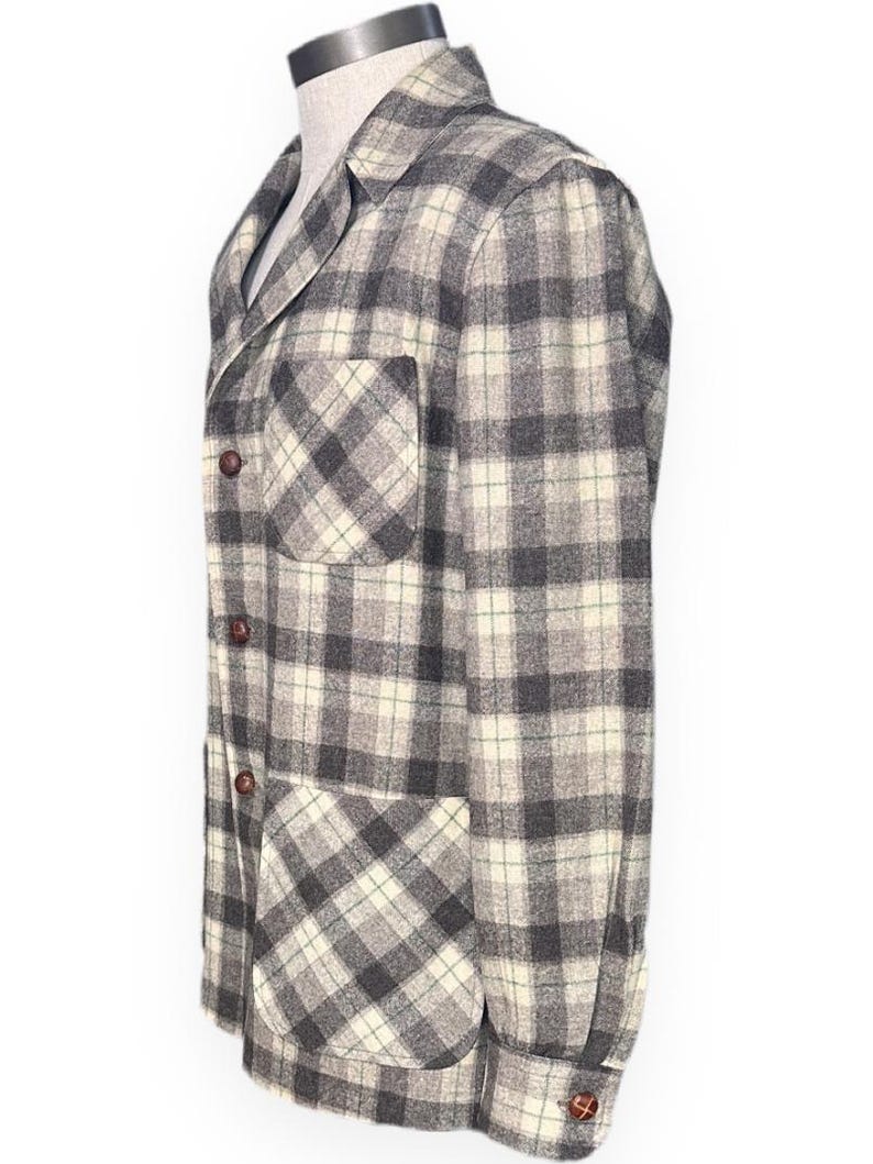 Puede incluir: Chaqueta a cuadros gris y crema con dos bolsillos delanteros y botones marrones. La chaqueta se exhibe en un maniqu&iacute; y tiene un dise&ntilde;o cl&aacute;sico y a medida. El patr&oacute;n de cuadros es una mezcla de gris, crema y un toque de verde.