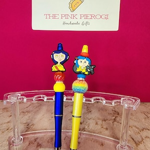 May include: Two decorative pens with character toppers, one blue and one yellow, displayed in a clear acrylic pen holder. The pens have colorful bead accents and are set against a pink background. The sign above reads "THE PINK PIEROGI Handmade Gifts."