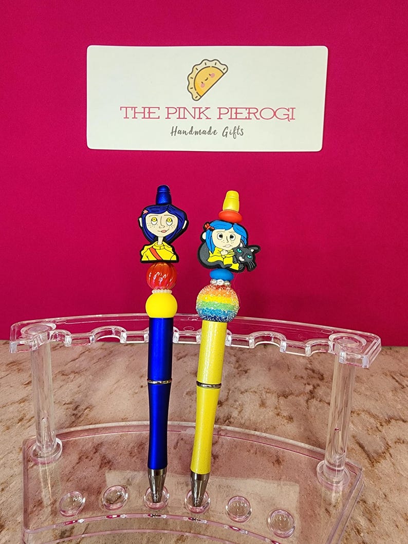 May include: Two decorative pens with character toppers, one blue and one yellow, displayed in a clear acrylic pen holder. The pens have colorful bead accents and are set against a pink background. The sign above reads "THE PINK PIEROGI Handmade Gifts."