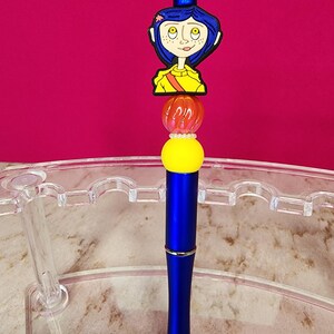 May include: A blue pen with a decorative topper featuring a cartoon character with blue hair and a yellow shirt. The pen has a yellow and orange bead design, and a silver tip. The background is a solid magenta color.