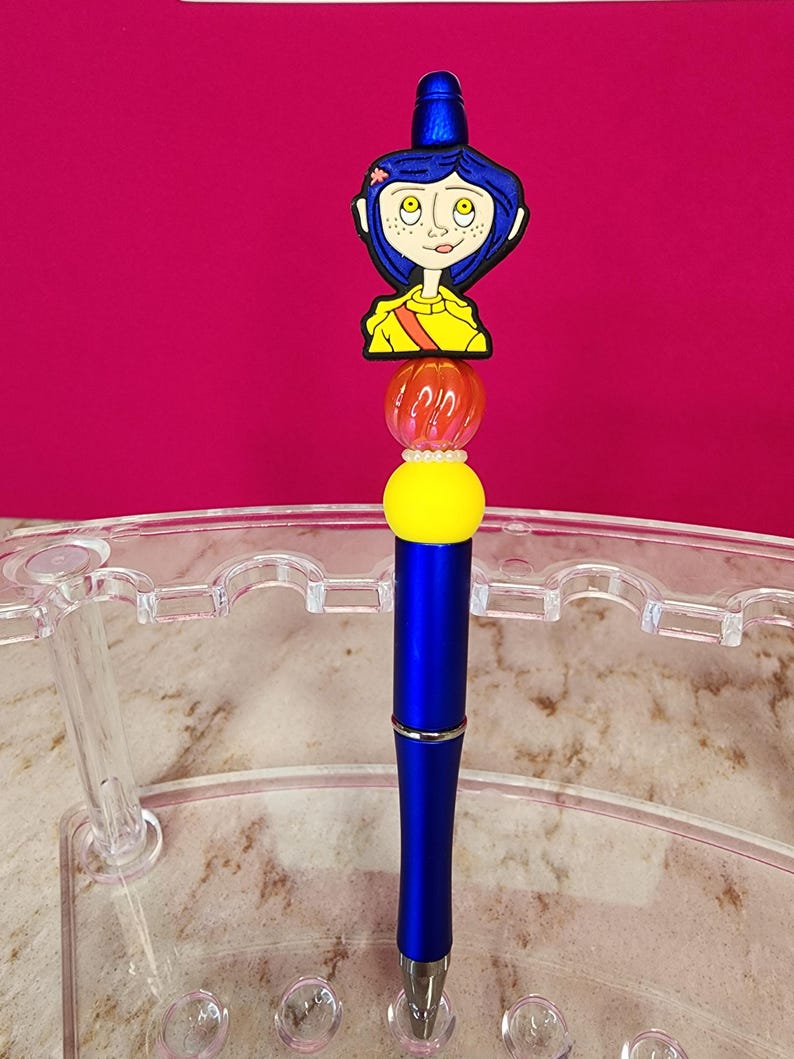 May include: A blue pen with a decorative topper featuring a cartoon character with blue hair and a yellow shirt. The pen has a yellow and orange bead design, and a silver tip. The background is a solid magenta color.