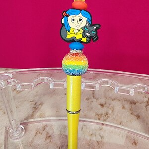 May include: A decorative pen with a yellow barrel and a rainbow-colored bead. The pen features a charm of a cartoon girl with blue hair and a black cat. The pen has a silver band and a yellow top.