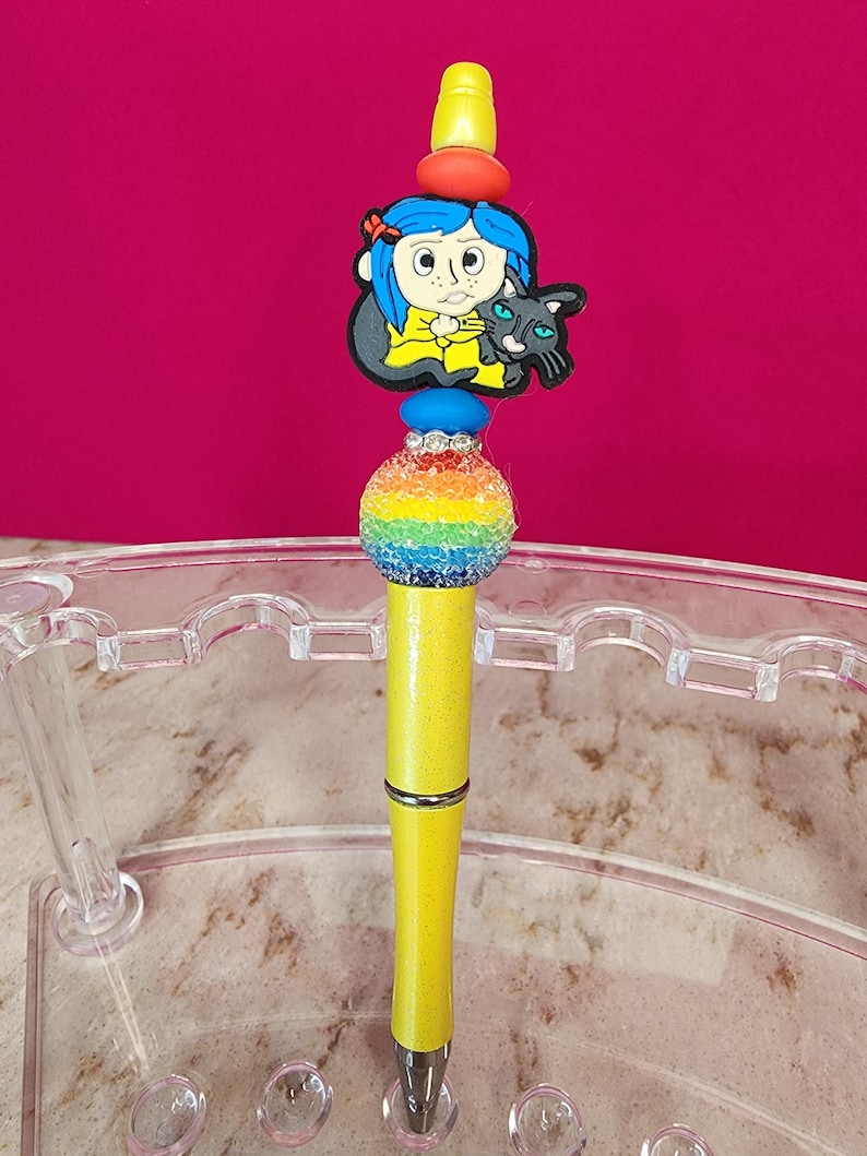 May include: A decorative pen with a yellow barrel and a rainbow-colored bead. The pen features a charm of a cartoon girl with blue hair and a black cat. The pen has a silver band and a yellow top.