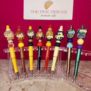 May include: A collection of decorative pens featuring character and house emblems from a popular fantasy series. The pens have colorful barrels and are displayed in a clear plastic stand. The text "THE PINK PIEROGI Handmade Gifts" is visible.
