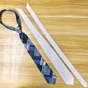 May include: A necktie featuring a patchwork pattern of various blue and white paisley and bandana-style prints, displayed alongside two long, thin, white rectangular strips of fabric that appear to be components for tie construction.
