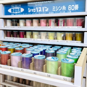 May include: A close-up of a shelf with various spools of thread in different colors. The spools are labeled with numbers and are organized in rows. The text on the sign above the shelf reads "フジックス シャッペンパン ミシン糸 60番".