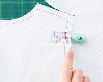 May include: A clear plastic ruler with pink markings is used to measure a white fabric pattern. The ruler has markings in inches and centimeters.