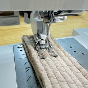 Könnte beinhalten: Nahaufnahme einer Nähmaschinennadel und eines Nähfußes, die einen beigefarbenen gesteppten Stoff nähen. Der Stoff wird durch die Maschine geführt und die Nadel bewegt sich auf und ab.