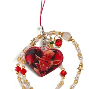 May include: A phone charm featuring a heart-shaped acrylic pendant with a red-toned character illustration, attached to a beaded loop. The strand includes clear, gold, and red beads, small gold star charms, and a small apple-shaped charm with a red stone.