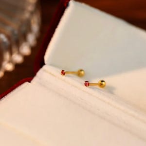 May include: Two gold-colored stud earrings with a small, round, red gemstone at the end of each post. The earrings are displayed on a white surface inside a red jewelry box. The earrings have a simple, elegant design.