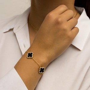 May include: Gold-toned bracelet featuring two black clover-shaped charms with a diamond-like border. The bracelet is connected by a delicate chain and is worn on a wrist. A matching gold necklace is also visible.