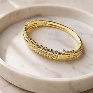 May include: A gold-toned hinged bangle bracelet featuring a central row of small, clear, sparkling stones flanked by two rows of textured, bead-like metal detailing. The bracelet includes a secure, integrated box clasp closure mechanism.