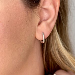 May include: A pair of silver hoop earrings with a slightly curved shape. The earrings are worn in the earlobe of a person with light brown hair.