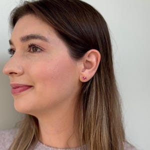 May include: A small heart-shaped stud earring featuring a vibrant pink gemstone set in a gold-toned metal frame. The earring is worn on the earlobe, showcasing a simple and compact design with a polished finish that catches the light.