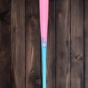 May include: A pink and blue baseball bat. The top portion is pink, transitioning to a light blue handle. A stylized design in blue is on the pink section. The bat is set against a dark wood grain background.