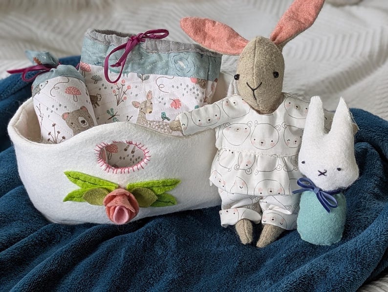 Peut inclure: Un panier en feutre blanc avec une application florale, contenant des articles en tissu imprim&eacute;s d'animaux de la for&ecirc;t. Un lapin en peluche v&ecirc;tu d'une tenue &agrave; motifs d'ours et un petit lapin blanc avec un n&oelig;ud bleu sont &eacute;galement pr&eacute;sents.