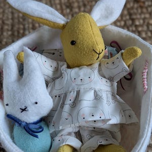 Peut inclure: Une poup&eacute;e lapin jaune faite &agrave; la main avec de longues oreilles et une tenue blanche et grise &agrave; motifs d'ours est assise dans un panier blanc. Une plus petite poup&eacute;e lapin blanche avec un n&oelig;ud bleu est &eacute;galement dans le panier.