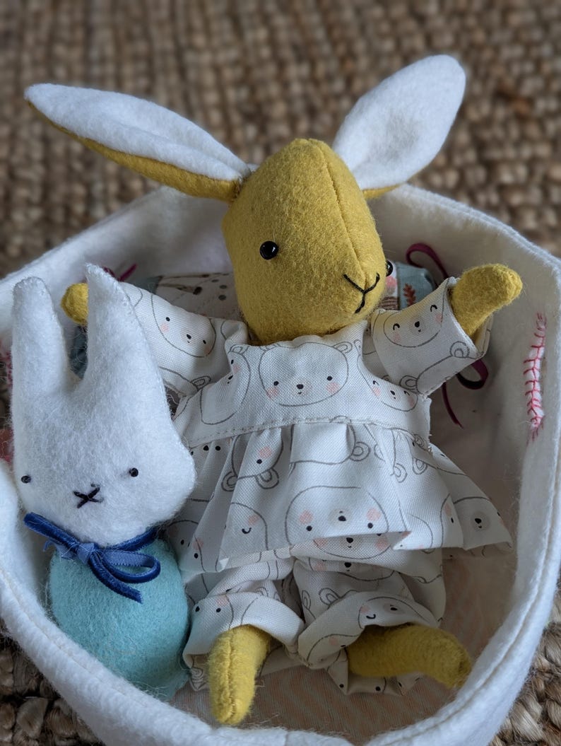 Peut inclure: Une poup&eacute;e lapin jaune faite &agrave; la main avec de longues oreilles et une tenue blanche et grise &agrave; motifs d'ours est assise dans un panier blanc. Une plus petite poup&eacute;e lapin blanche avec un n&oelig;ud bleu est &eacute;galement dans le panier.