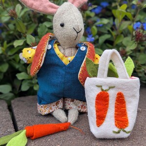 May include: A handmade stuffed bunny doll with pink ears, wearing a blue denim overall and a yellow patterned shirt. It holds a white felt bag with two orange carrot appliqués and a felt carrot. The bunny is a whimsical, handcrafted item.