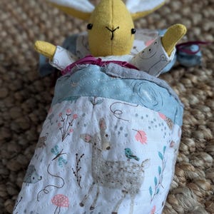 Peut inclure: Une peluche faite &agrave; la main avec une t&ecirc;te et des bras de lapin jaunes, nich&eacute;e dans un sac de couchage &agrave; motifs bleu et blanc. Le sac de couchage pr&eacute;sente une sc&egrave;ne de for&ecirc;t avec un cerf, des oiseaux et des fleurs.