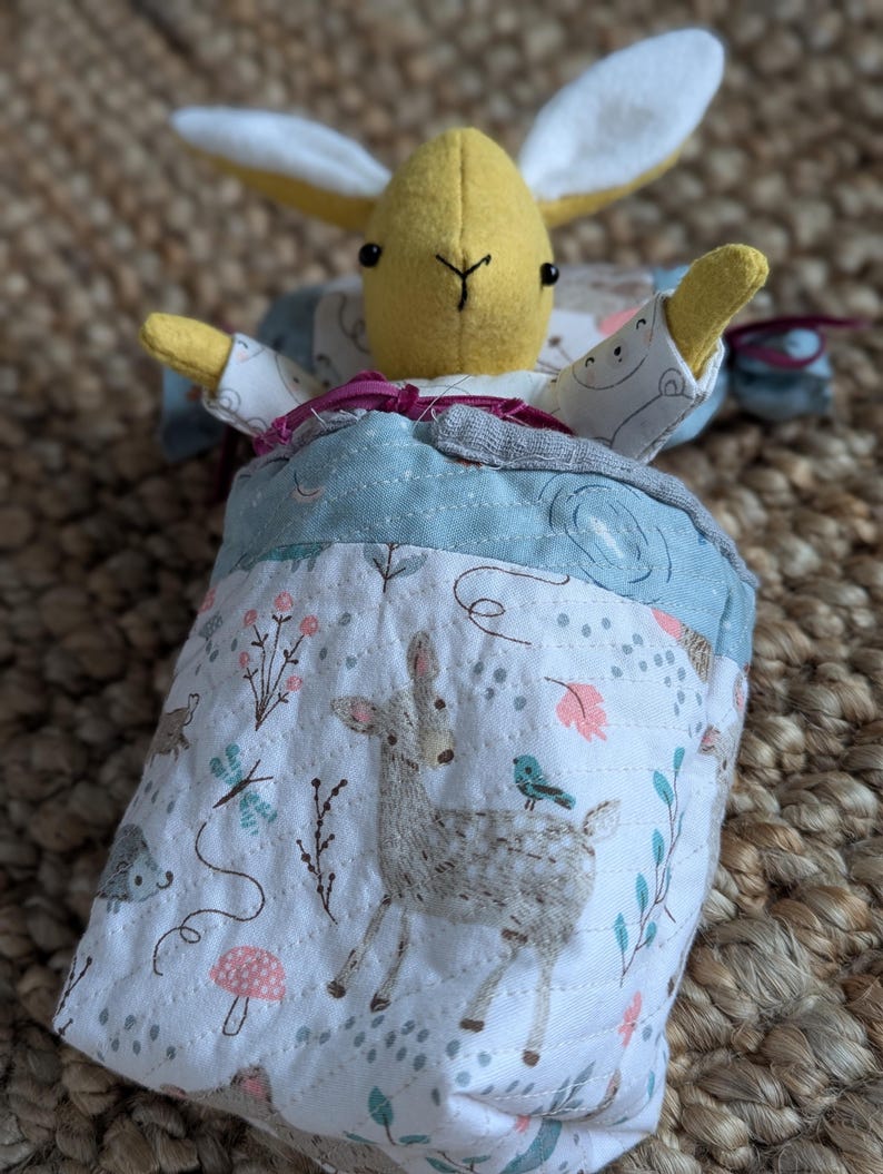 Peut inclure: Une peluche faite &agrave; la main avec une t&ecirc;te et des bras de lapin jaunes, nich&eacute;e dans un sac de couchage &agrave; motifs bleu et blanc. Le sac de couchage pr&eacute;sente une sc&egrave;ne de for&ecirc;t avec un cerf, des oiseaux et des fleurs.