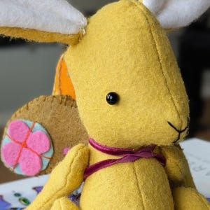 Könnte beinhalten: Ein handgemachter gelber Plüschhase mit weißen Innenohren und einem rosa Blumendetail an der Seite. Der Hase hat ein schwarzes Knopfauge und ein lila Band um den Hals. Das ist ein Stofftier.