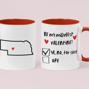 Puede incluir: Taza de café blanca con borde y asa rojos. La taza tiene un contorno negro del estado de Nebraska con un corazón rojo en su interior. El otro lado de la taza dice "Be my midwest valentine?" con una casilla de verificación junto a "Ya, no, for sure" y una casilla de verificación vacía junto a "Ope".