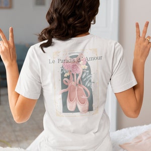 Puede incluir: Camiseta blanca con un estampado gráfico de zapatillas de ballet rosas y flores. El texto "Le Paradis d'Amour" está impreso en la camiseta.