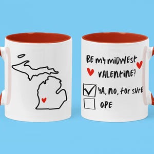 Puede incluir: Taza de cerámica blanca con borde y asa rojos. La taza tiene un contorno negro del estado de Michigan con un corazón rojo en el centro. El otro lado de la taza dice "Be my midwest valentine?" con una casilla de verificación y las palabras "Ya, no, for sure" y "Ope" escritas debajo.