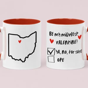 Puede incluir: Taza de cerámica blanca con borde y asa rojos. La taza presenta un contorno negro del estado de Ohio con un corazón rojo en su interior. El otro lado de la taza dice "Be my midwest valentine?" con una casilla de verificación y las opciones "Ya, no, for sure" y "Ope".