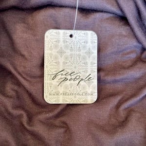 May include: Close-up of a tag from Free People, featuring the brand's logo in cursive script. The tag is rectangular with rounded corners and a decorative floral pattern. The background is a textured, dark purple fabric.
