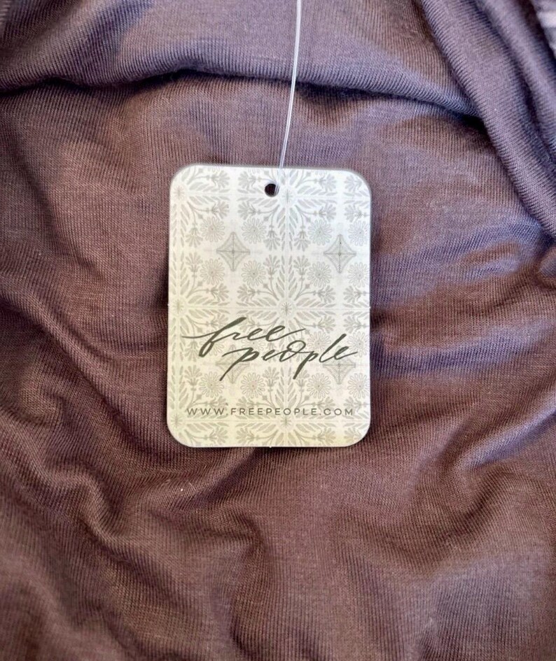 May include: Close-up of a tag from Free People, featuring the brand's logo in cursive script. The tag is rectangular with rounded corners and a decorative floral pattern. The background is a textured, dark purple fabric.