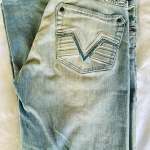 Peut inclure: Jean en denim bleu clair délavé. La poche arrière présente un design unique avec des coutures bleues. Un patch en cuir marron avec le texte "Affliction 1979 Black Premium" est visible à la taille.