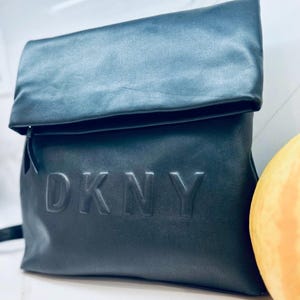 May include: Black DKNY backpack with a folded-over top. The bag features the embossed DKNY logo on the front. The backpack is made of a smooth, matte material and has a simple, modern design. The bag is shown against a white background.