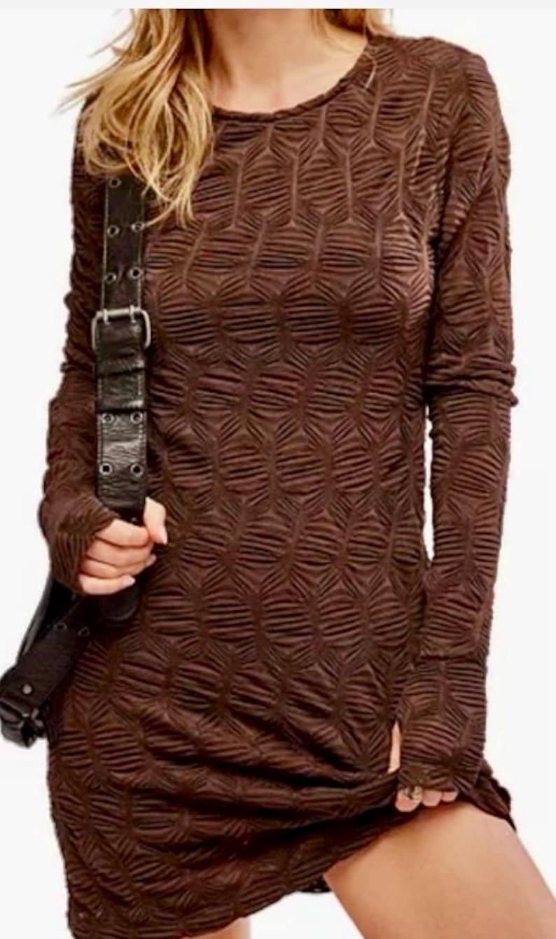 May include: A long-sleeved, brown dress with a textured, geometric pattern. The dress has a crew neck and fitted sleeves. A black leather shoulder bag with a silver buckle is visible. The dress is knee-length and has a slit on the side.