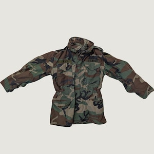 May include: A long-sleeve field jacket featuring a woodland camouflage pattern in shades of brown, green, and black. The jacket includes a high collar, two chest pockets with flaps, and the text U.S. ARMY embroidered above the right chest pocket.