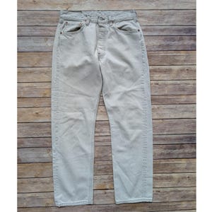 Puede incluir: Un par de pantalones vaqueros de color beige claro con un diseño clásico de cinco bolsillos, cierre de botones y remaches metálicos de color cobre en las esquinas de los bolsillos. Corte recto con costuras visibles.