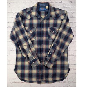 May include: A long-sleeve button-down shirt featuring a plaid pattern in shades of navy blue, cream, and grey. The shirt includes a pointed collar, two chest pockets with snap closures, and metal snap buttons down the front and on the cuffs.