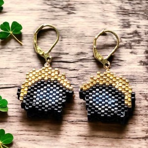 May include: A pair of gold and black beaded earrings with a blue and white pixelated design. The earrings are dangle style with a gold hook.