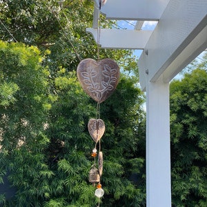 May include: A wooden heart-shaped wind chime with carved leaf details hangs from a white pergola. The chime features multiple small bells and clear glass beads.