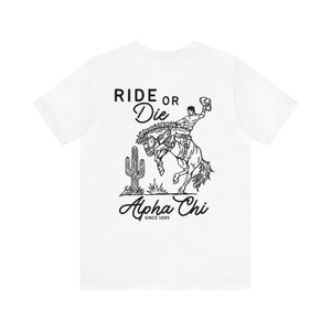 Può includere: Maglietta bianca con un'illustrazione in bianco e nero di un cowboy che cavalca un cavallo selvaggio. Il testo "Ride or Die" è sopra l'illustrazione e "Alpha Chi" è sotto l'illustrazione con il testo "Since 1885" sotto.