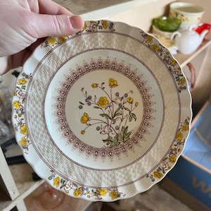 May include: A white ceramic plate with a scalloped edge, featuring a floral design with yellow flowers and green leaves. The plate has a brown and cream-colored border with a woven pattern and a decorative inner ring.
