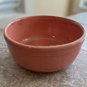 May include: A pink ceramic bowl with a smooth, glossy finish. The bowl has a simple, rounded shape and a slightly flared rim.