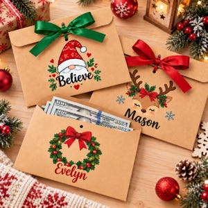 Könnte beinhalten: Drei braune Kraftpapier-Umschläge mit weihnachtlichen Motiven. Einer zeigt einen Wichtel und das Wort Believe mit einer grünen Schleife. Ein weiterer zeigt ein Rentier und den Namen Mason mit einer roten Schleife. Der dritte zeigt einen Kranz.