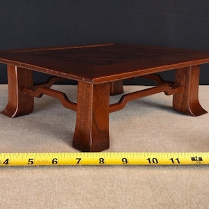 May include: A small, square wooden table with a dark brown finish. The table has a decorative base with curved supports and measures approximately 12 inches wide. The table is designed for display or decorative purposes.