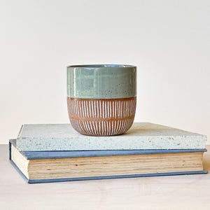 May include: A handcrafted ceramic mug with a two-tone design. The upper section is a speckled sage green, and the lower section is a warm brown with vertical white stripes. The mug rests on two books.