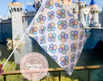 May include: A small white flag with a pattern of colorful puzzle pieces, held in front of a castle. The flag has a white pole and a circular sticker that reads "Courage + Kind". The flag is a stroller flag.