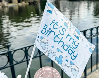 May include: A white stroller flag with the text "it's my Birthday" in blue, along with Mickey Mouse balloon graphics. The flag is held by a white clip. The flag is a Courage + Kind product.