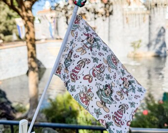 May include: A small white flag with a festive pattern featuring Disney characters in holiday attire. The flag is held by a white plastic clip and topped with a Mickey Mouse figure. The background includes a castle and a body of water.