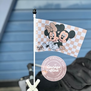 May include: A stroller flag featuring a checkered pattern in white and tan, with a graphic of Minnie and Mickey Mouse. The flag is attached to a white pole with a black topper and a white clip. The flag also has the text "Courage + Kind".